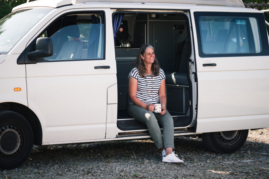 Photograph of Claire seated in the entrance door of her white campervan holding a cup of tea and smiling